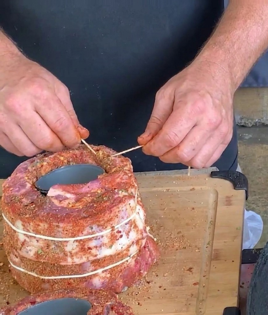 Hands using white butchers twine and a toothpick to tie and secure a large cylindrical roll of seasoned pork ribs wrapped around a can.
