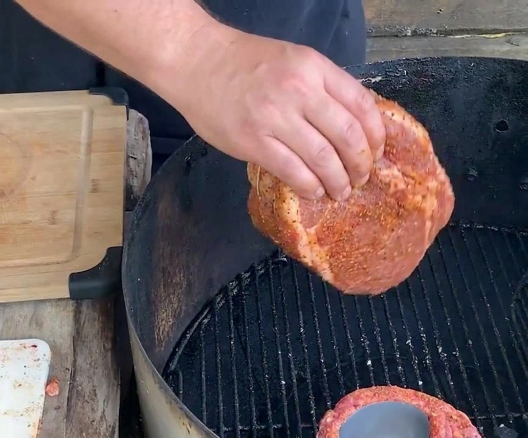 A hand lowering a tied, seasoned cylindrical rib cup onto a black steel charcoal grill grate for slow roasting.
