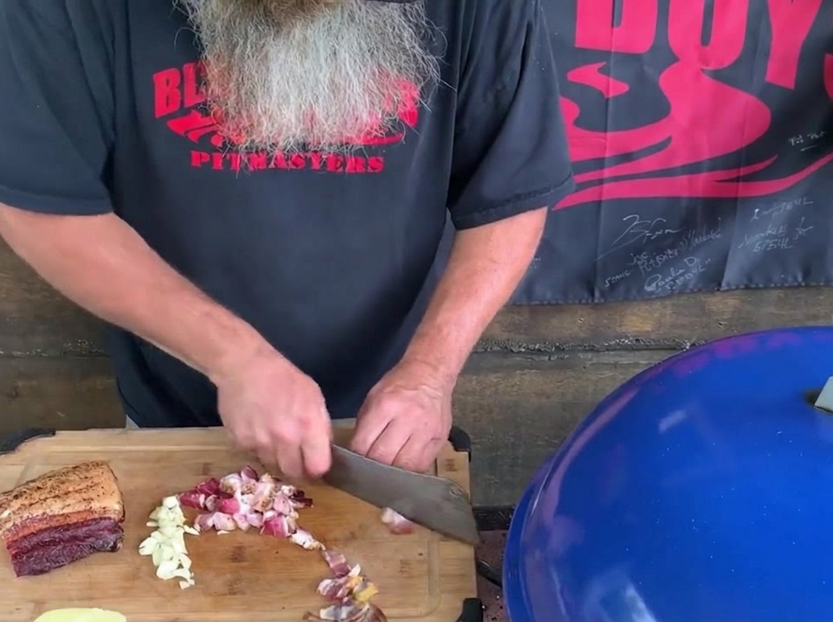 A man with a long gray beard using a large kitchen cleaver to chop bacon bits and garlic cloves on a wooden cutting board next to a blue grill.