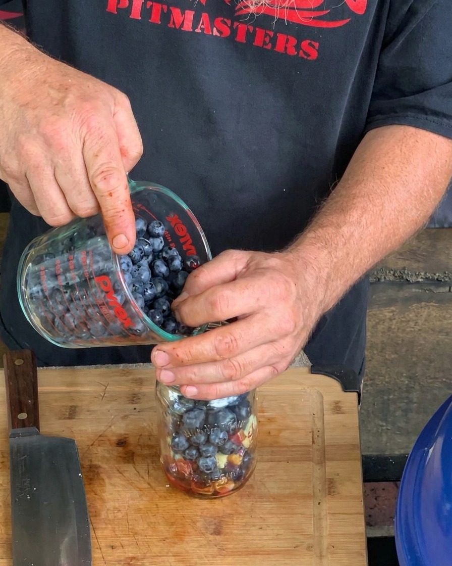 A close-up of hands pouring fresh blueberries from a Pyrex measuring cup into a glass mason jar containing other liquid and solid sauce ingredients.