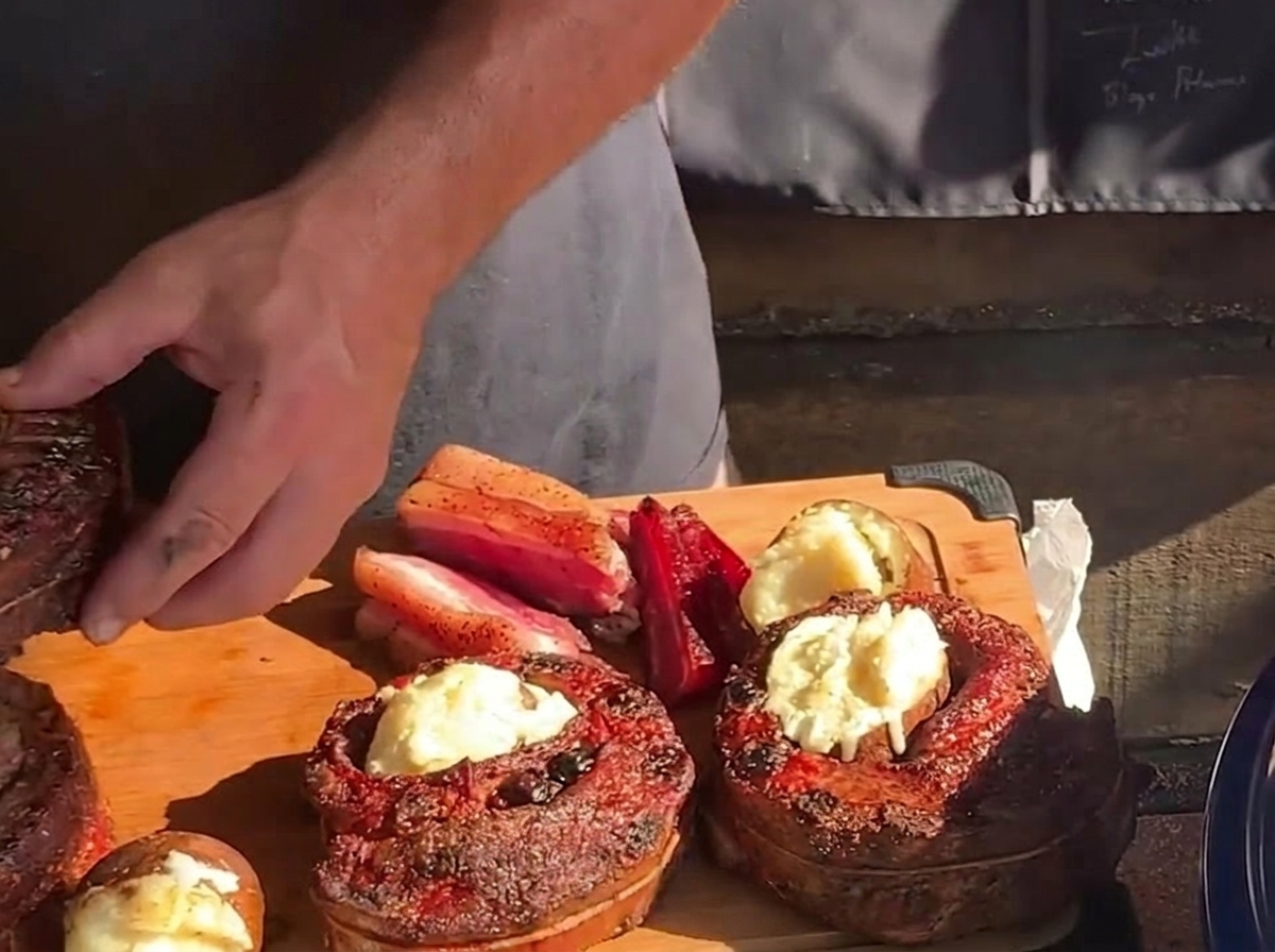 A hand placing a hot, buttered roasted potato into the hollow center of a cooked, round rib cup on a wooden cutting board.