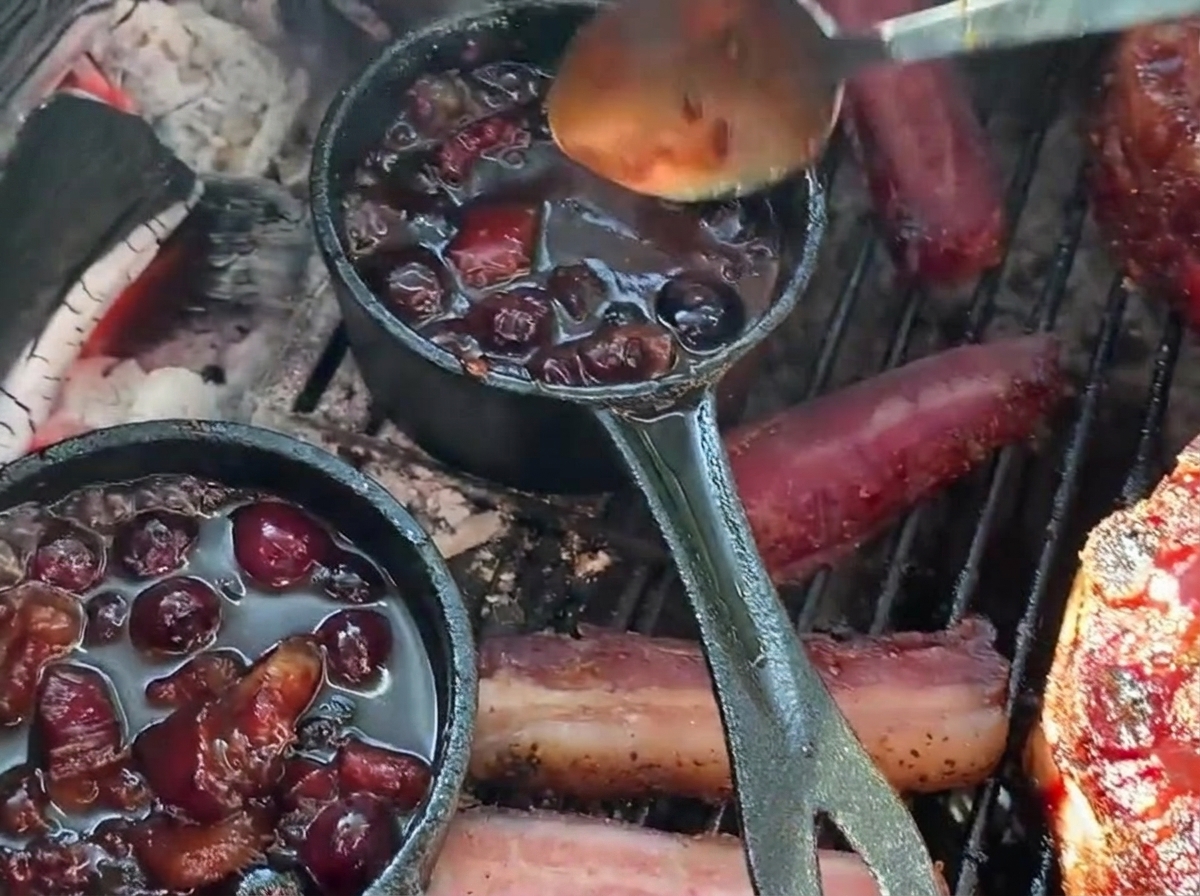 Spooning rich blueberry barbecue sauce from a small cast iron pan over a stuffed rib cup on the grill.