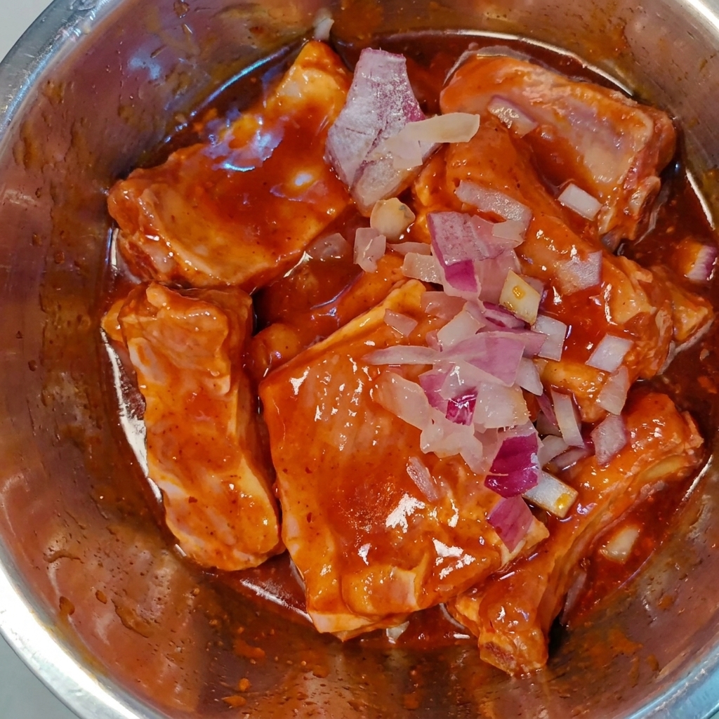 Finely chopped red onions sprinkled over marinated raw pork ribs in a metal bowl.