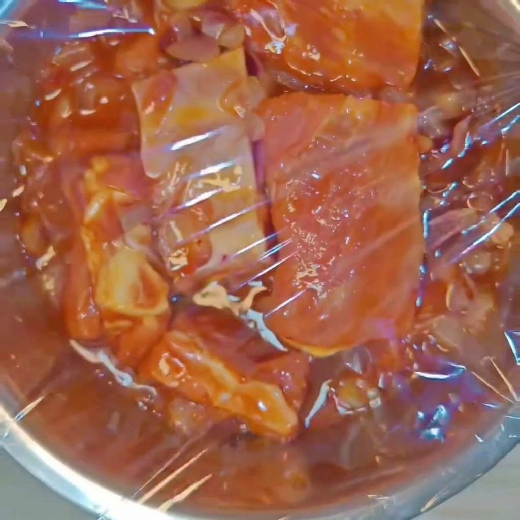 A metal bowl containing seasoned pork ribs tightly sealed with clear plastic wrap.