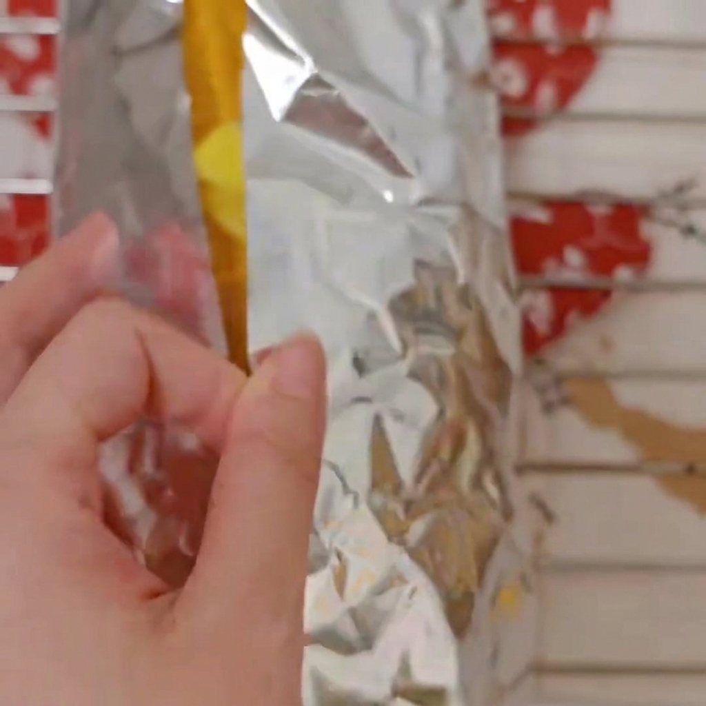 Fingers folding and wrapping sweet corn cobs inside a sheet of silver aluminum foil.