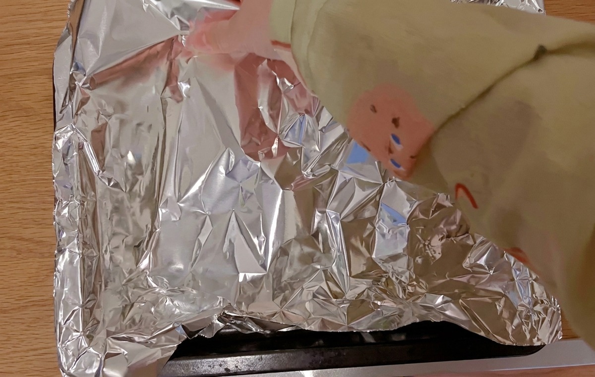 Hands covering marinated pork ribs on a black baking tray with a sheet of shiny aluminum foil.