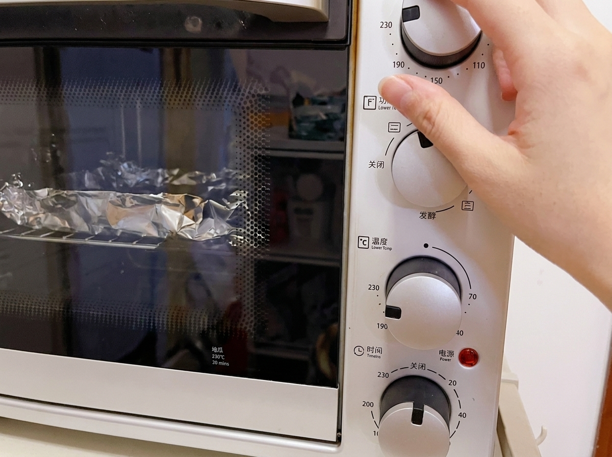 Close-up of a hand adjusting the temperature dial of a white electric countertop oven to 230°C, with foil-wrapped items visible on the wire rack inside.