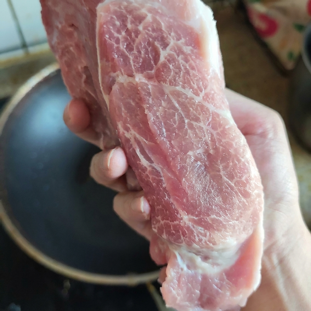A raw piece of well-marbled pork collar meat held up by hand to exhibit its fresh texture and fat distribution.