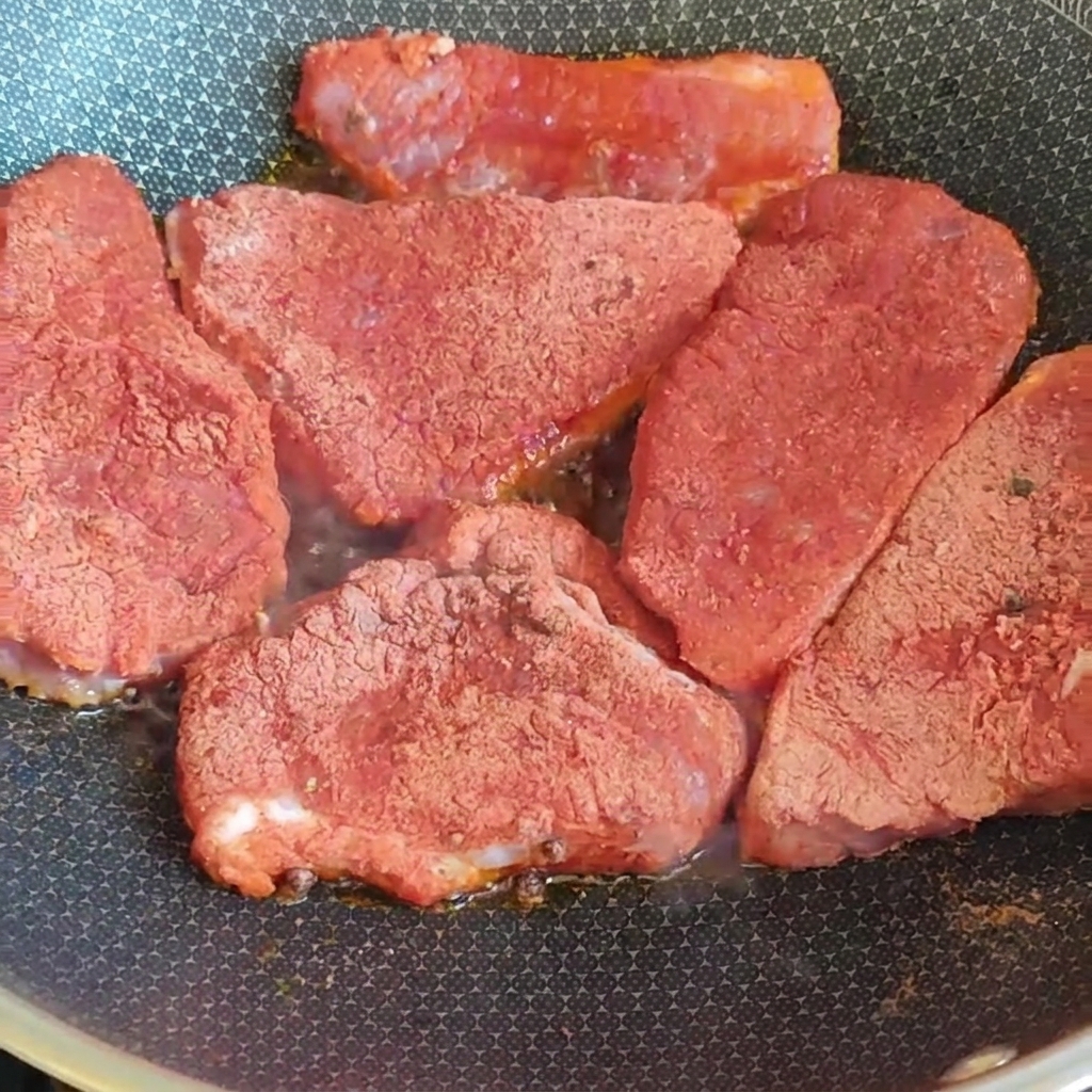 Thick pork slices covered in a red spice mixture searing inside a hot pan on the stove.