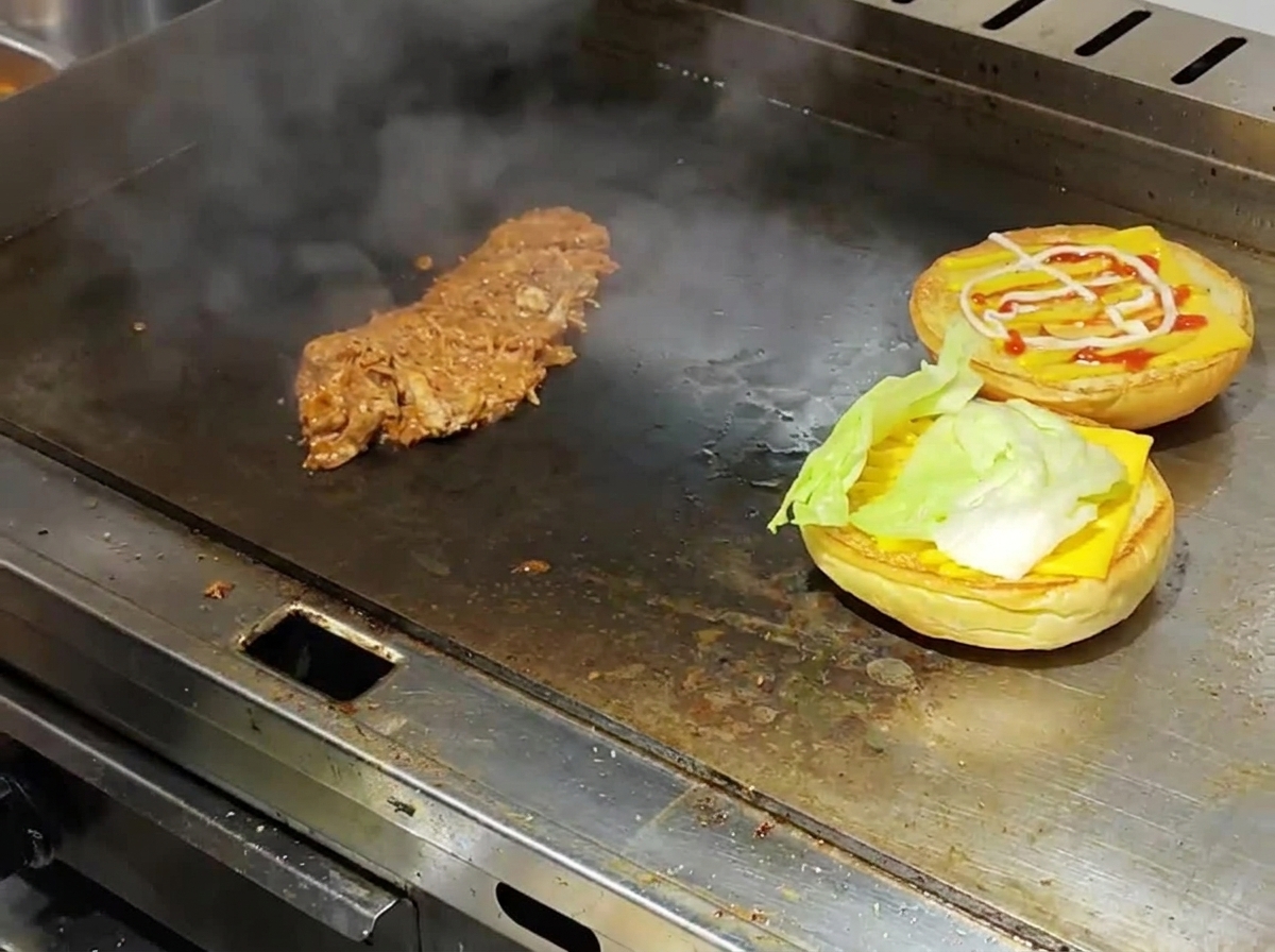 Toasted burger buns layered with cheese and lettuce alongside hot shredded pulled pork on a steaming commercial griddle.