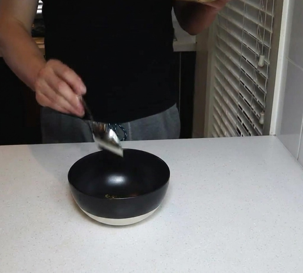 A person holding a jar and using a spoon to add dry spice powders into a black bowl on a white countertop.