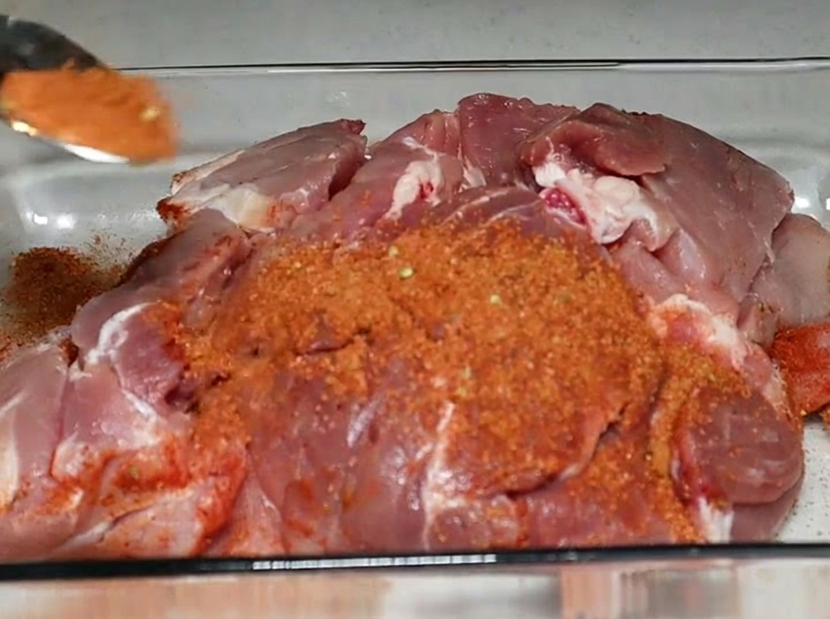 A spoonful of reddish spice rub being sprinkled onto a large cut of raw pork shoulder sitting in a glass baking dish.