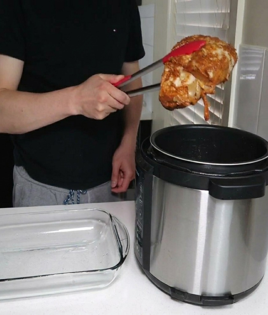 A person using red-tipped metal tongs to lift a large piece of cooked pork shoulder out of an electric pressure cooker next to an empty glass dish.