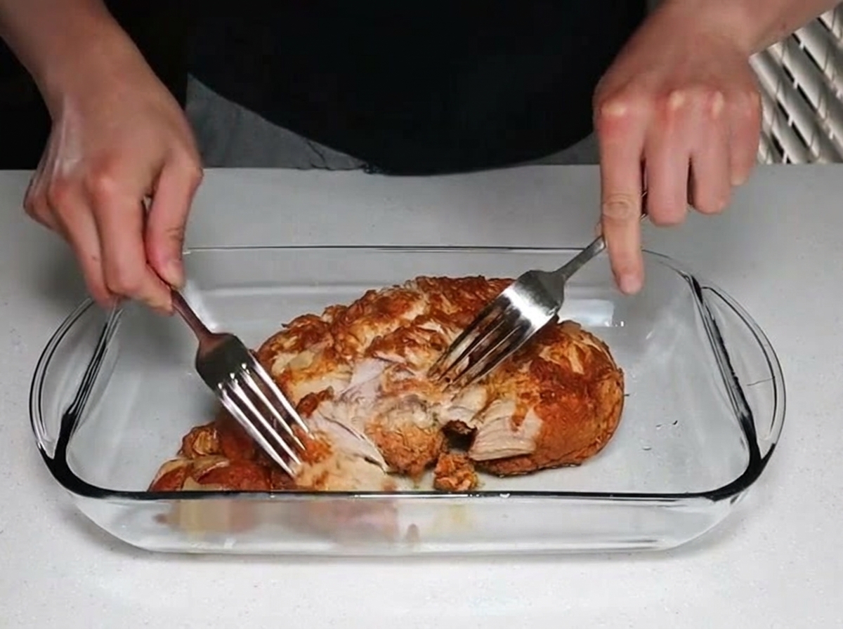 Two hands using stainless steel forks to pull apart and shred a large piece of tender cooked pork inside a clear glass baking dish.
