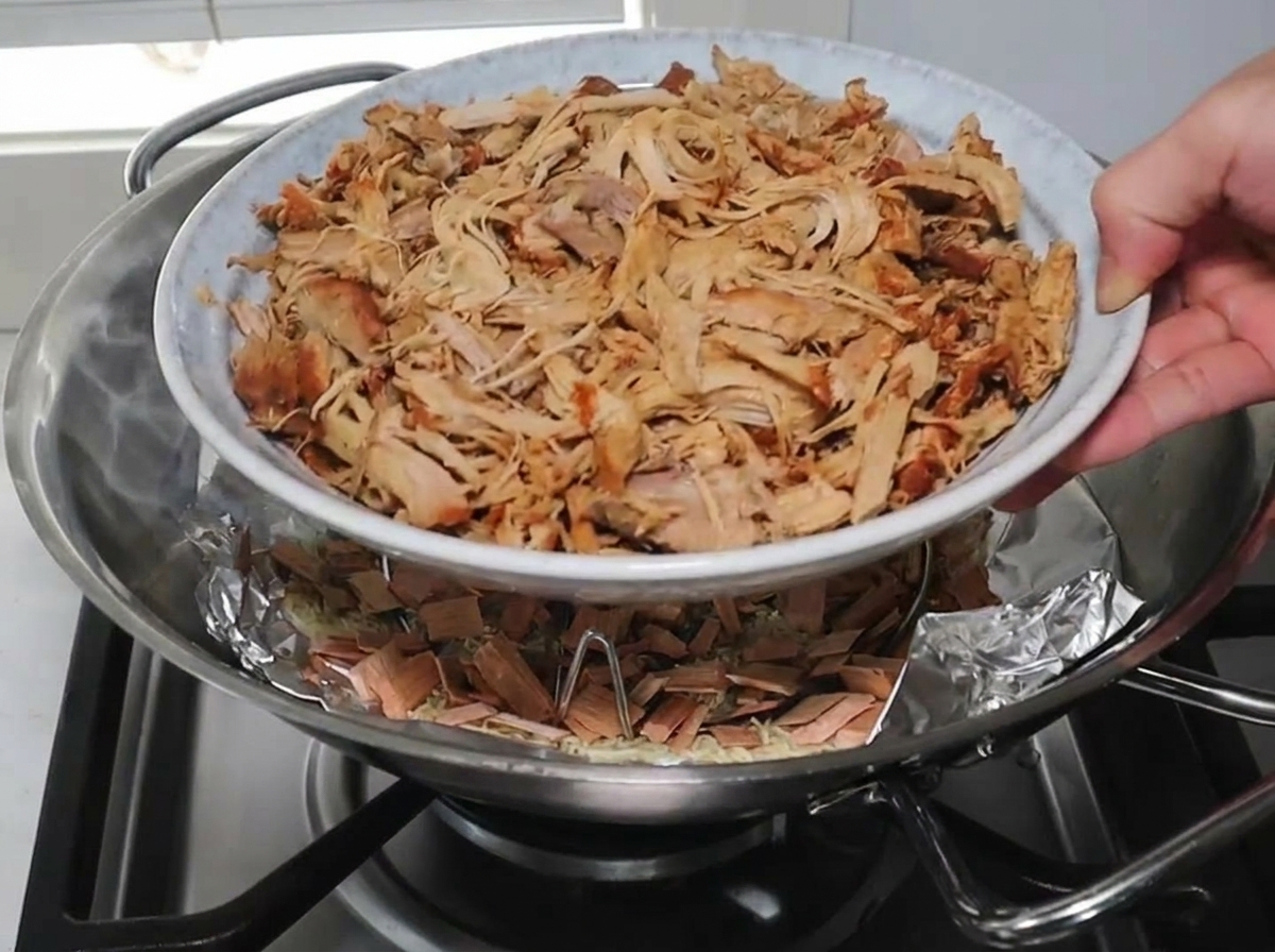 A hand lowering a white plate piled high with shredded pork onto a wire rack inside a wok over smoking wood chips.
