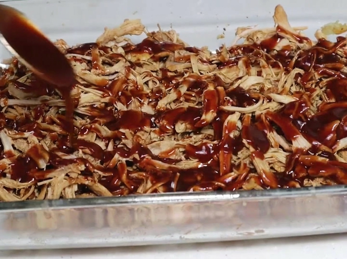 A spoonful of thick, dark barbecue sauce being drizzled over a layer of smoked shredded pork in a clear glass baking dish.