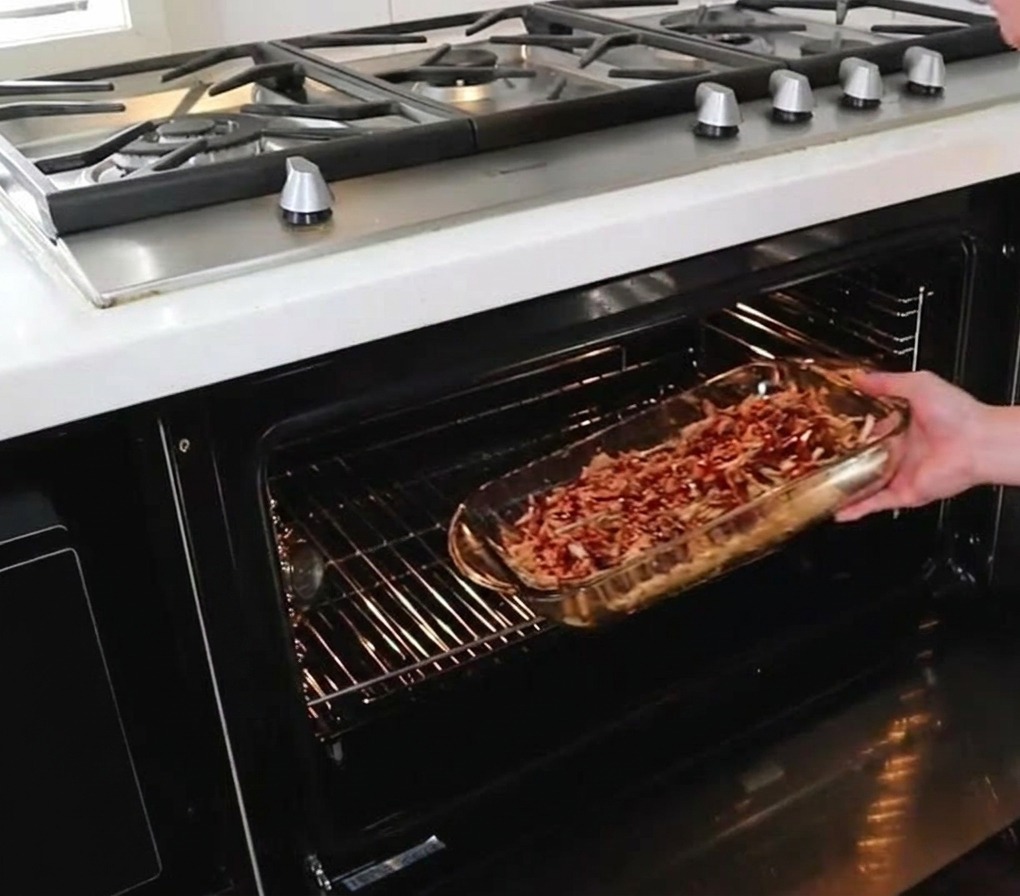 A person sliding a glass baking dish filled with sauced pulled pork onto the middle rack of a hot oven.