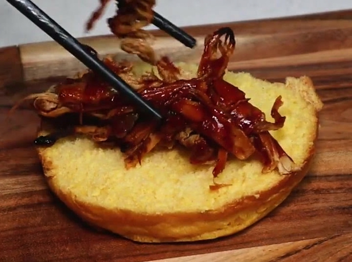 A pair of chopsticks placing succulent shredded pulled pork coated in rich, dark barbecue sauce onto the golden surface of a toasted burger bun half.