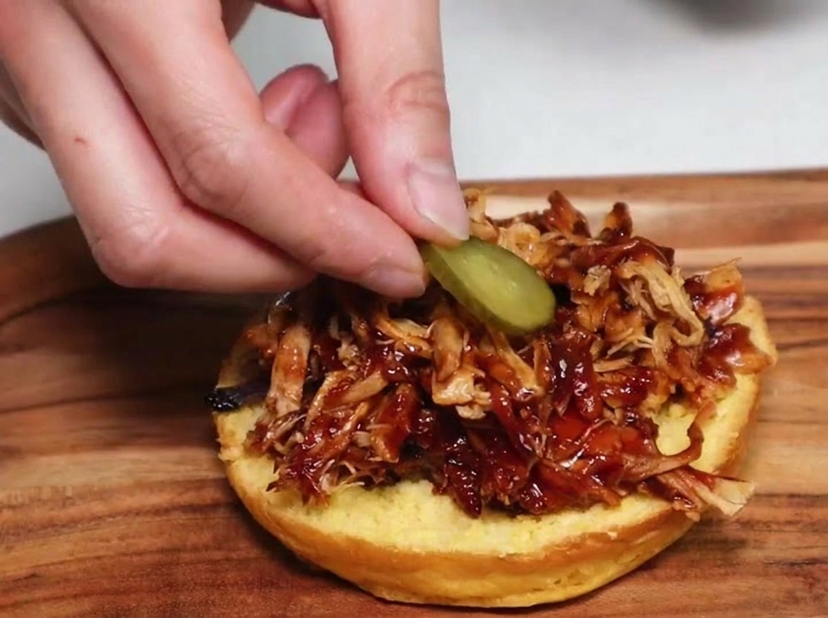 A hand placing a round pickle slice onto a pile of shredded barbecue pulled pork on a toasted burger bun.