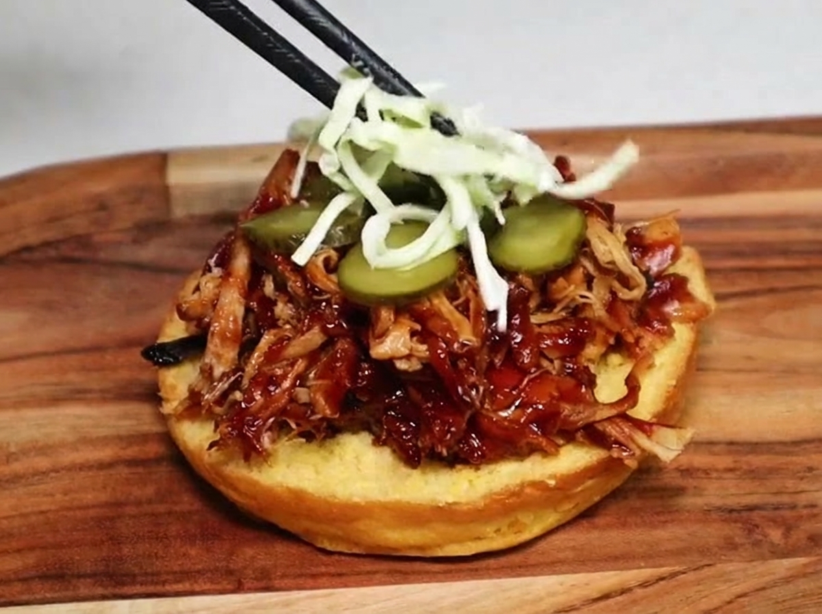 Chopsticks placing shredded green cabbage coleslaw on top of pickle slices and pulled pork inside a burger bun.