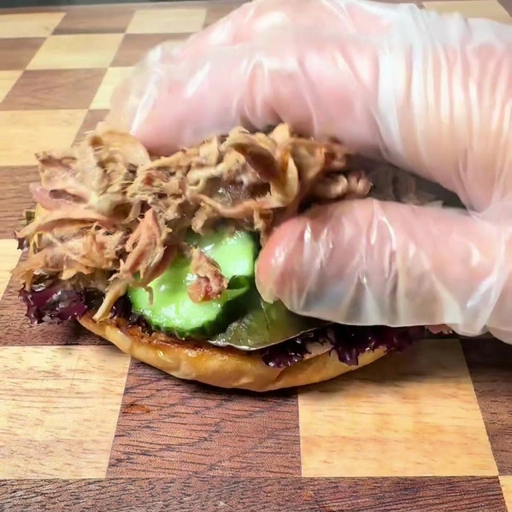 A hand wearing a clear plastic glove placing tender, shredded pulled pork onto a burger base with lettuce and cucumber slices.