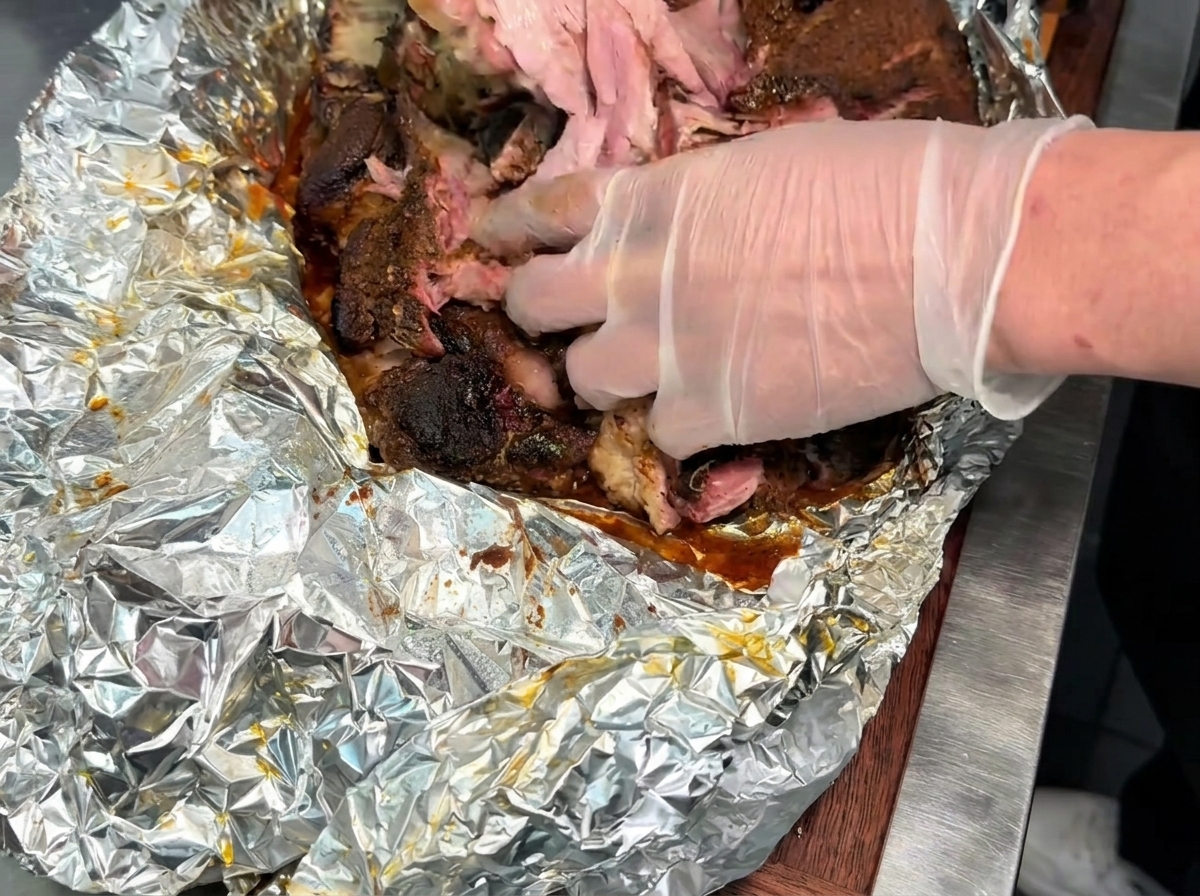 Gloved hands pulling apart and shredding a large piece of tender, cooked smoked pork on a foil-lined surface.