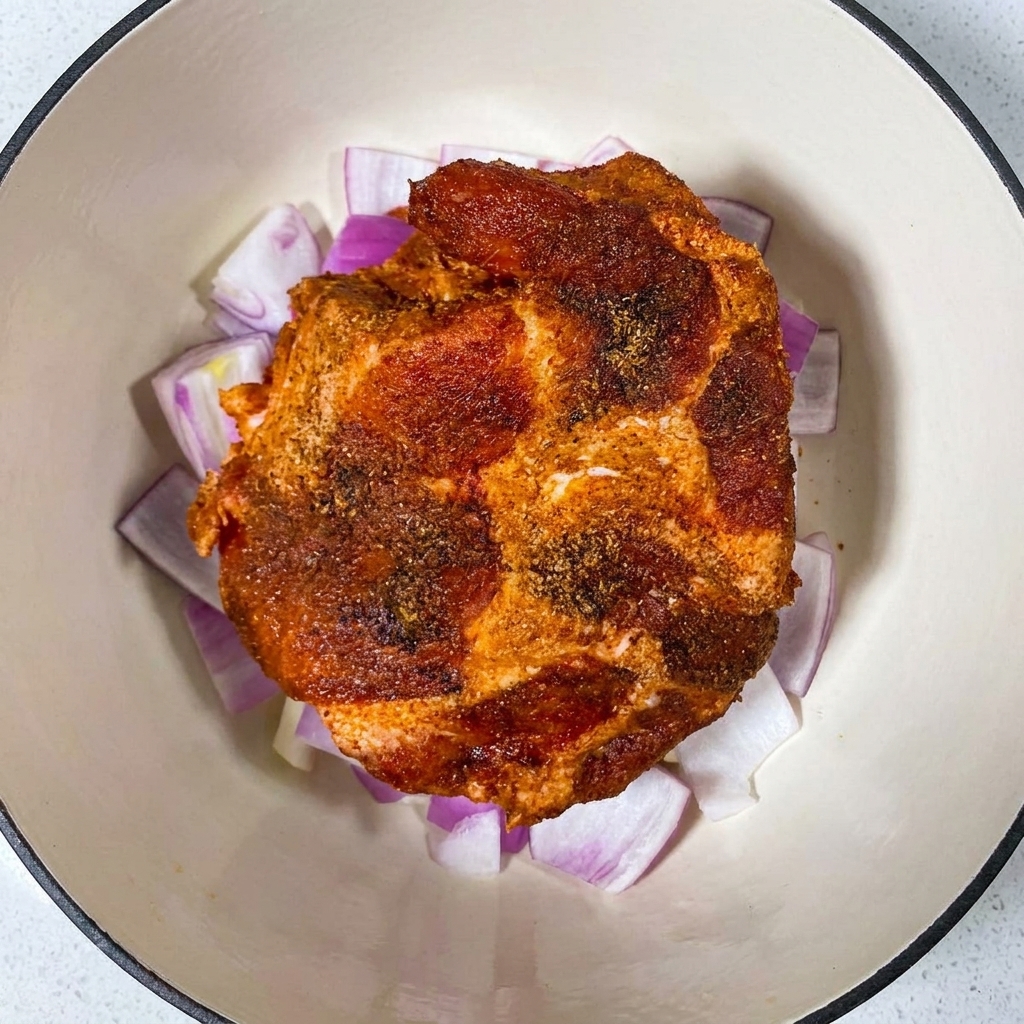An overhead view of a spice-rubbed pork shoulder resting on top of chopped red onions inside a white enameled pot.