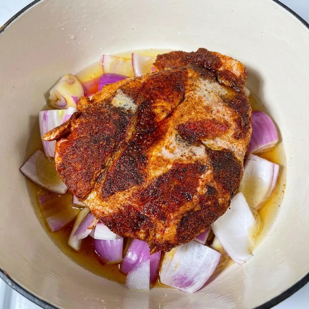 Braising liquids and beer poured into a pot surrounding a seasoned pork shoulder on a bed of red onions.