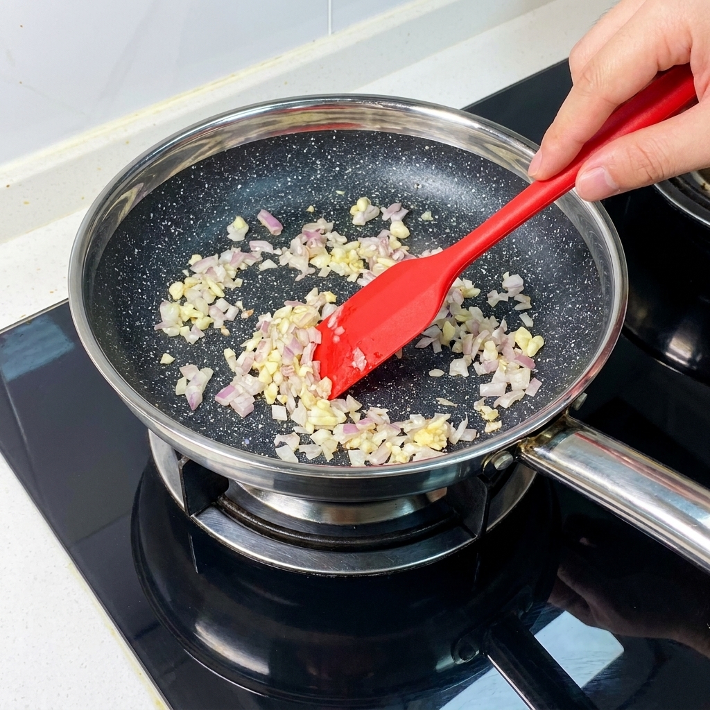 Finely diced onions and minced garlic being sautéed in a frying pan using a red spatula.