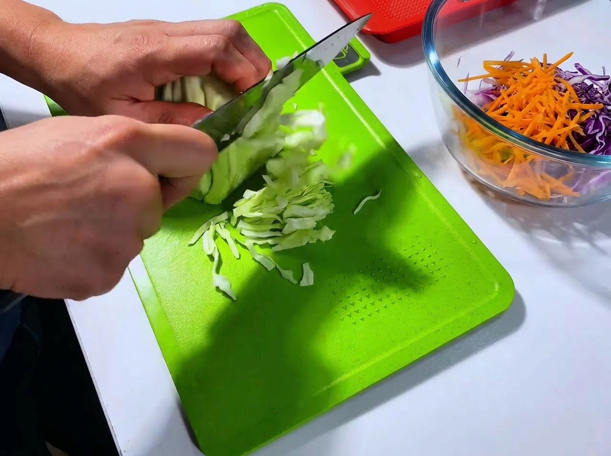 A chef slicing a head of green cabbage into thin shreds with a large knife on a bright green cutting board.