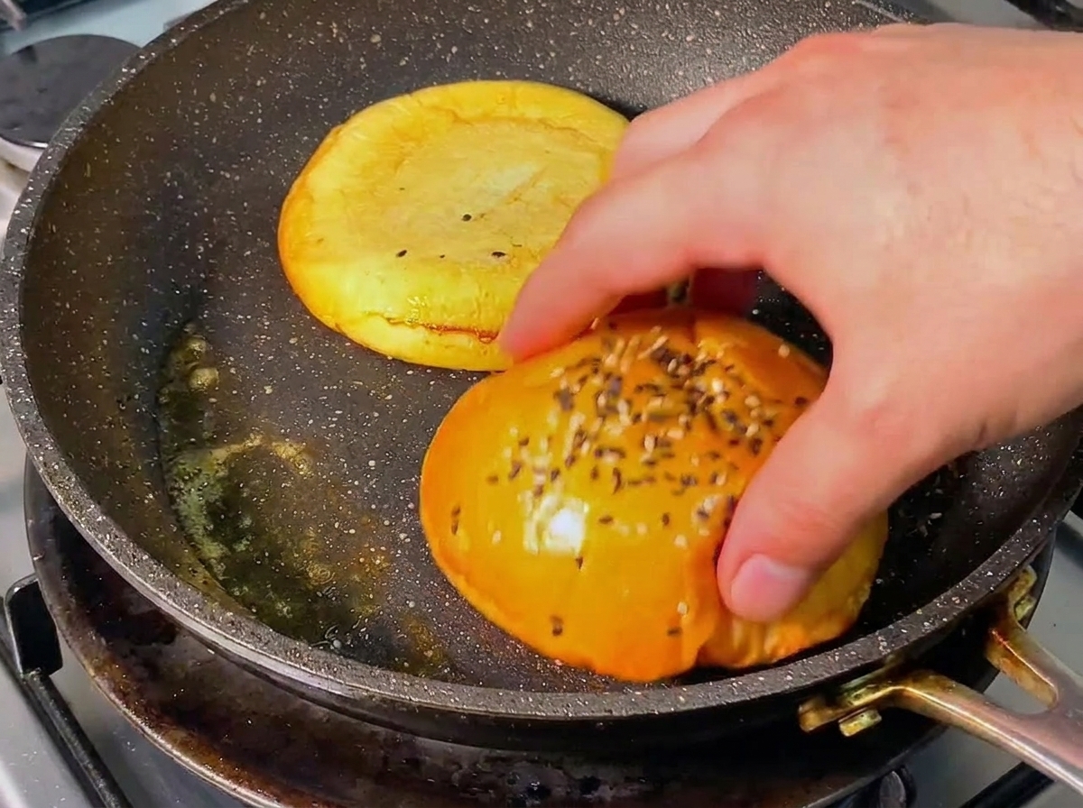 A hand turning a golden-brown burger bun half in a seasoned skillet with melted butter.
