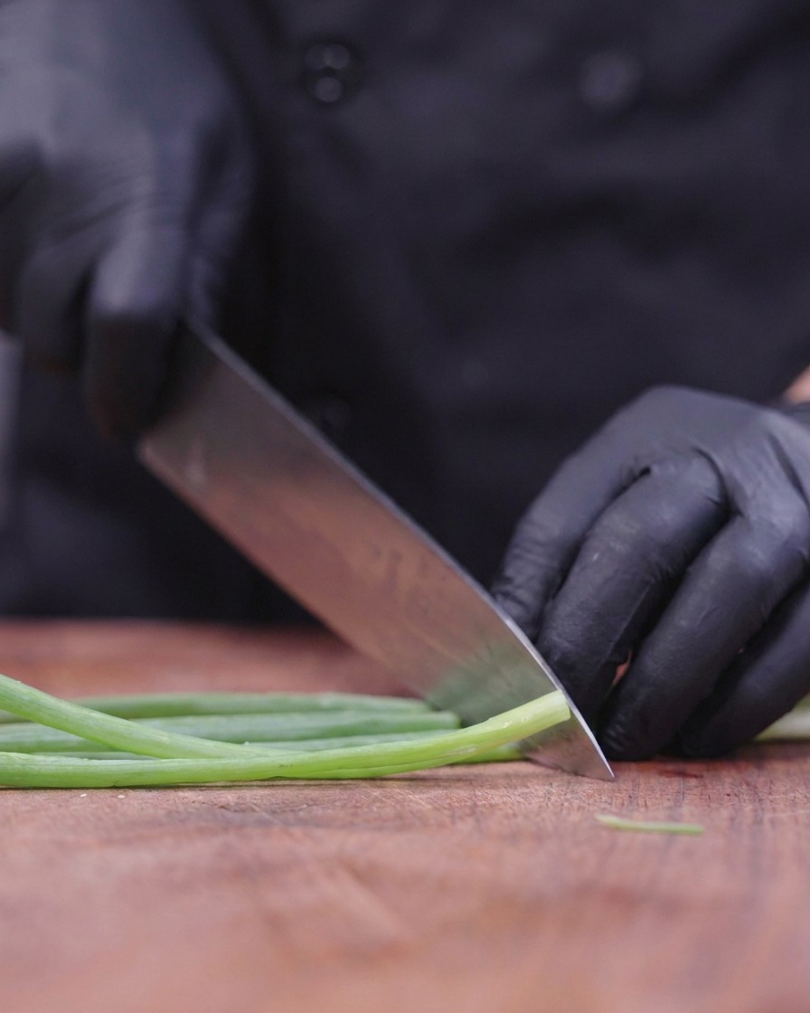 A chef wearing black gloves using a large knife to finely slice green scallion stalks on a wooden board.