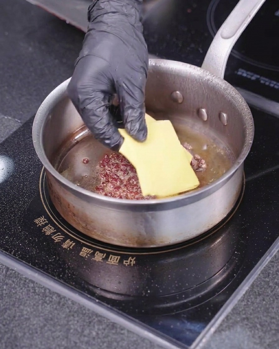 A gloved hand placing a square slice of yellow cheese onto a cooking meat patty inside a hot skillet.