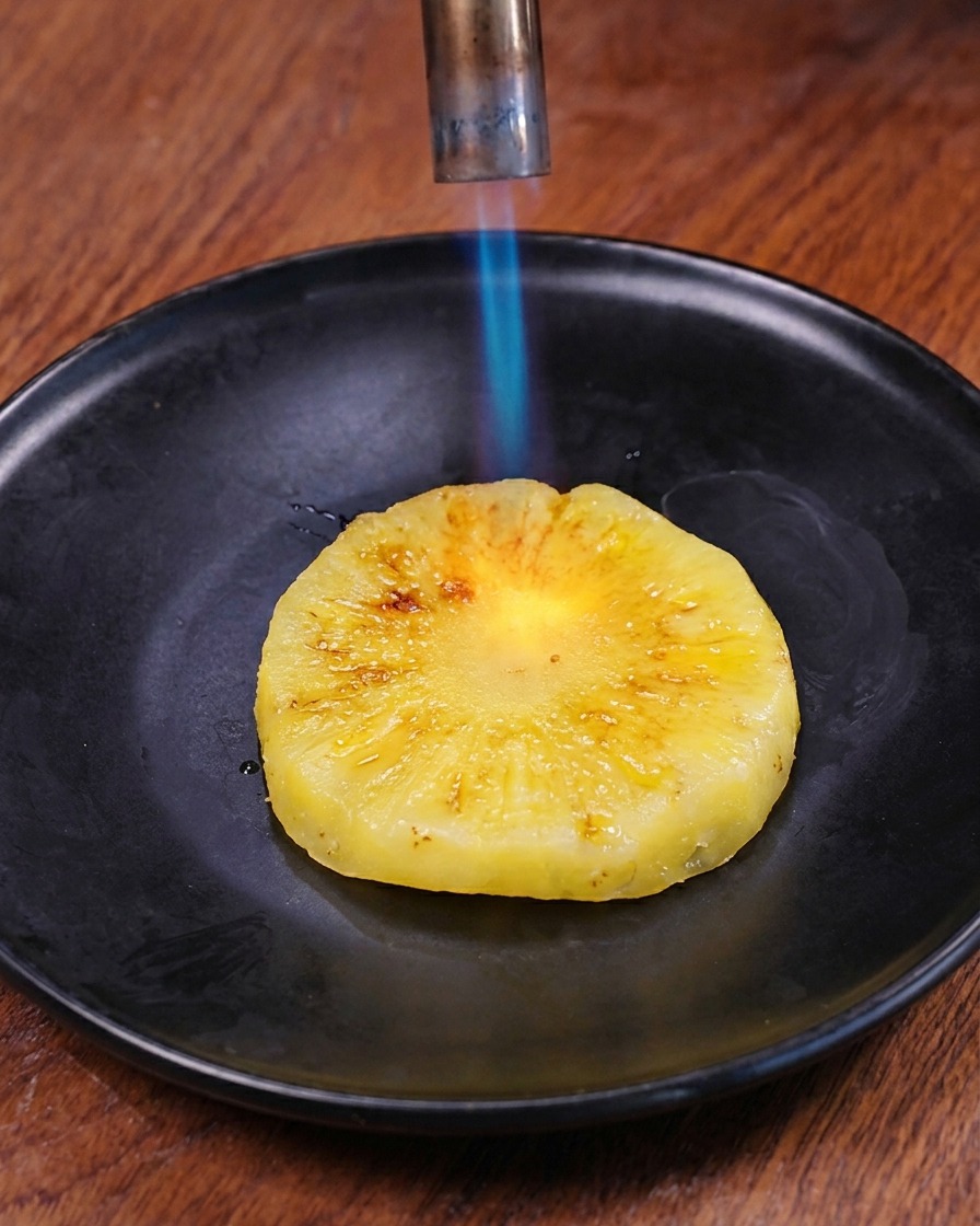 A blue flame from a kitchen blowtorch charring the surface of a fresh pineapple ring on a dark plate.