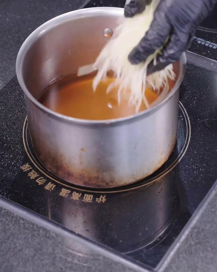 A hand wearing a black glove dropping finely shredded white potatoes into a saucepan filled with hot cooking oil.