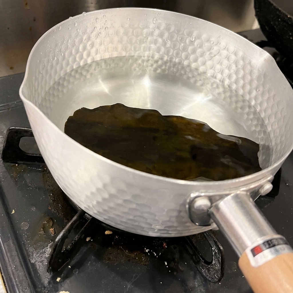 A single piece of dark, dried kelp rests at the bottom of a hammered aluminum pot filled with clear, cold water.