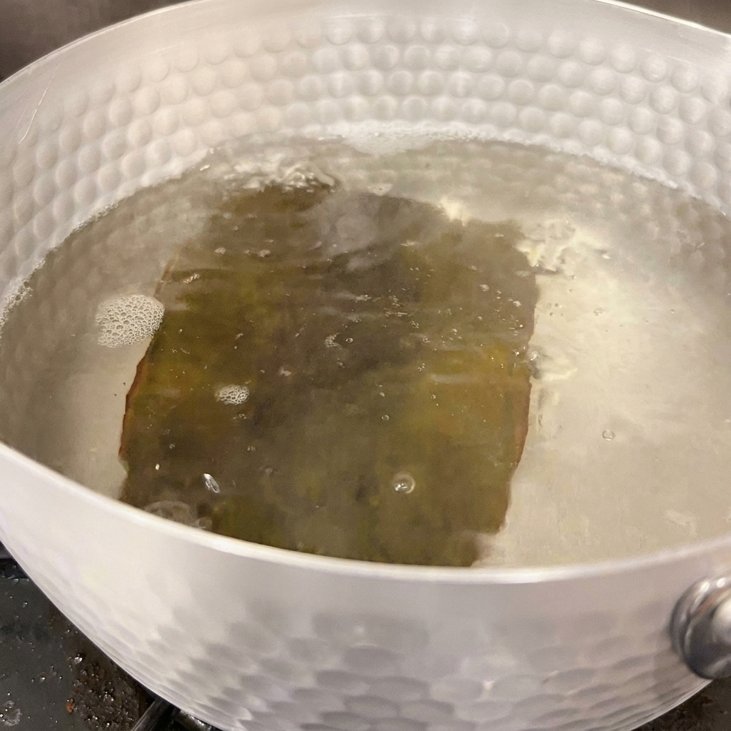 Water bubbling gently around a piece of rehydrated kelp inside a silver pot on the stove.