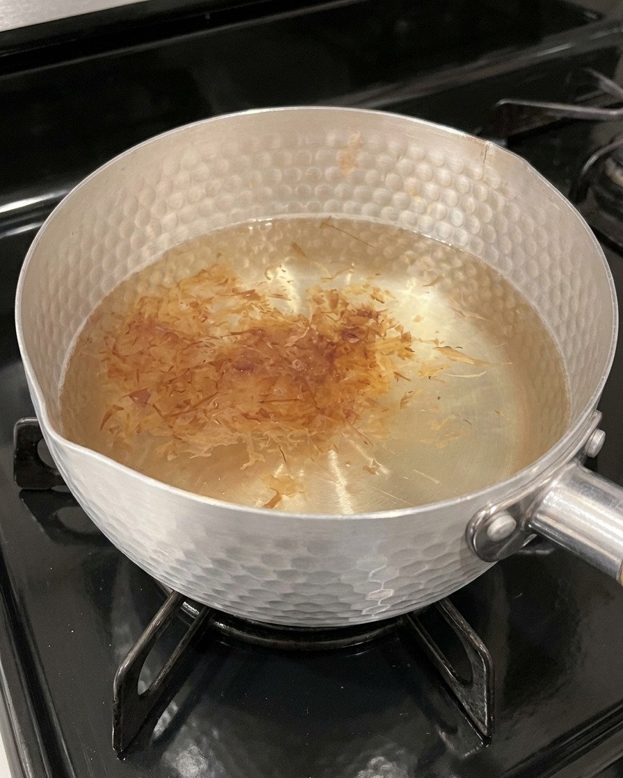 Light brown, papery bonito flakes steeping on the surface of hot, amber-tinted broth in a pot.