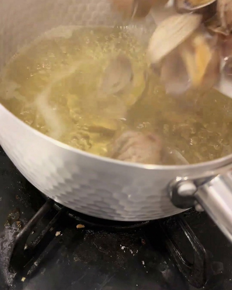 Fresh clams being dropped into a boiling, clear dashi stock inside a metallic pot on a gas stove.