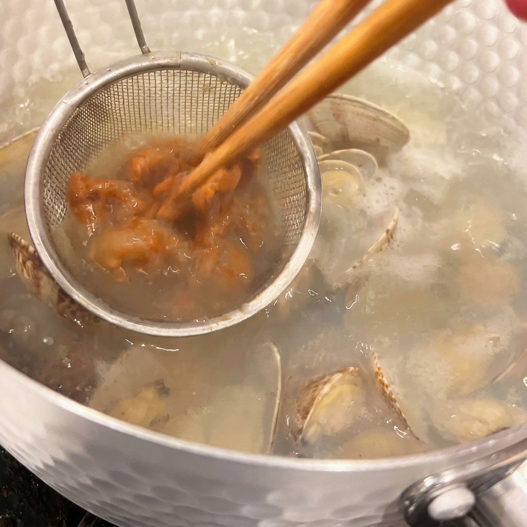 A small handheld strainer holding savory brown miso paste while chopsticks blend it into a hot clam soup.
