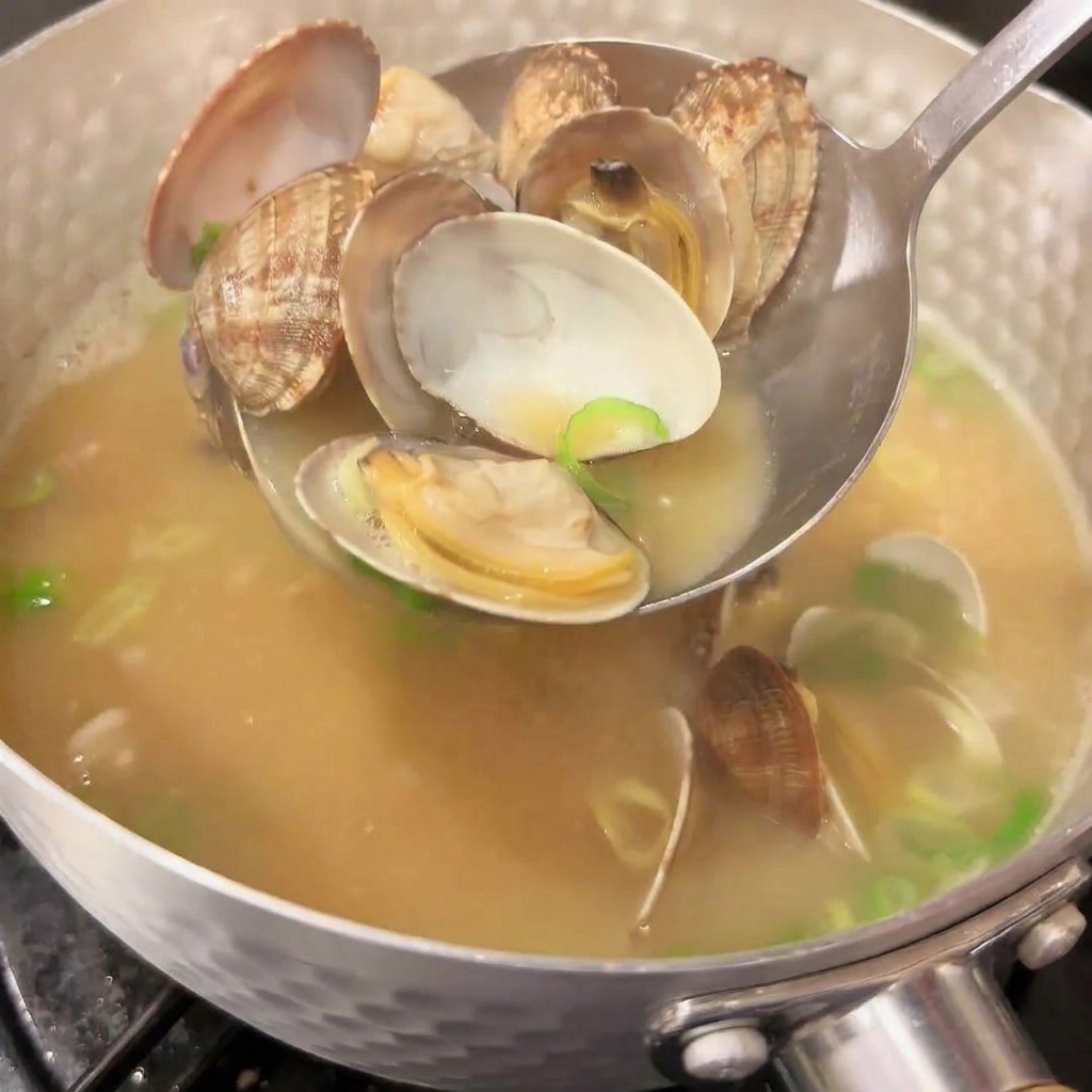 A metal ladle scooping up opened clams from a pot of steaming hot Japanese miso soup, with vibrant chopped green onions mixed into the broth.