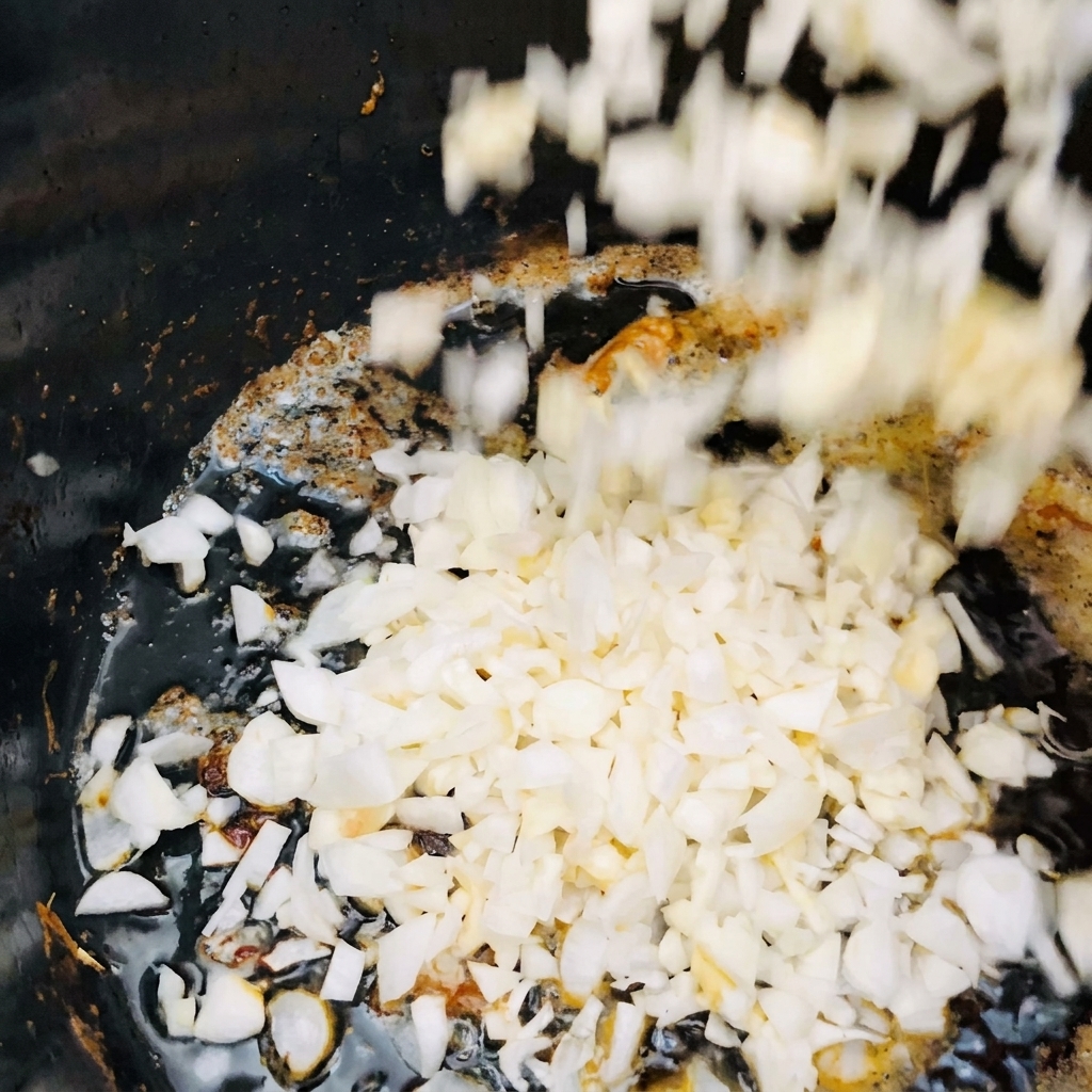 Diced white onions and minced garlic falling into a dark pot with shimmering hot oil.