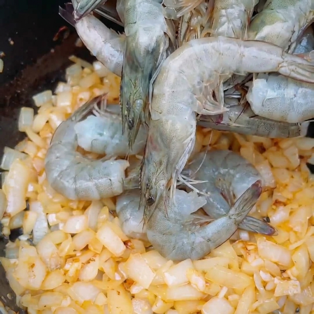 Fresh grey whole shrimp being placed onto a bed of sautéed diced onions in a pot.
