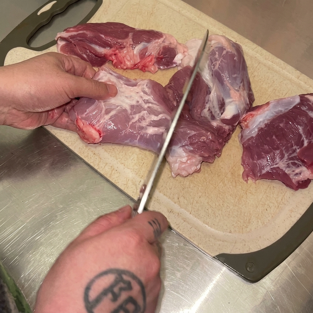 Hands using a knife to cut raw pork hind leg into chunks on a cutting board.