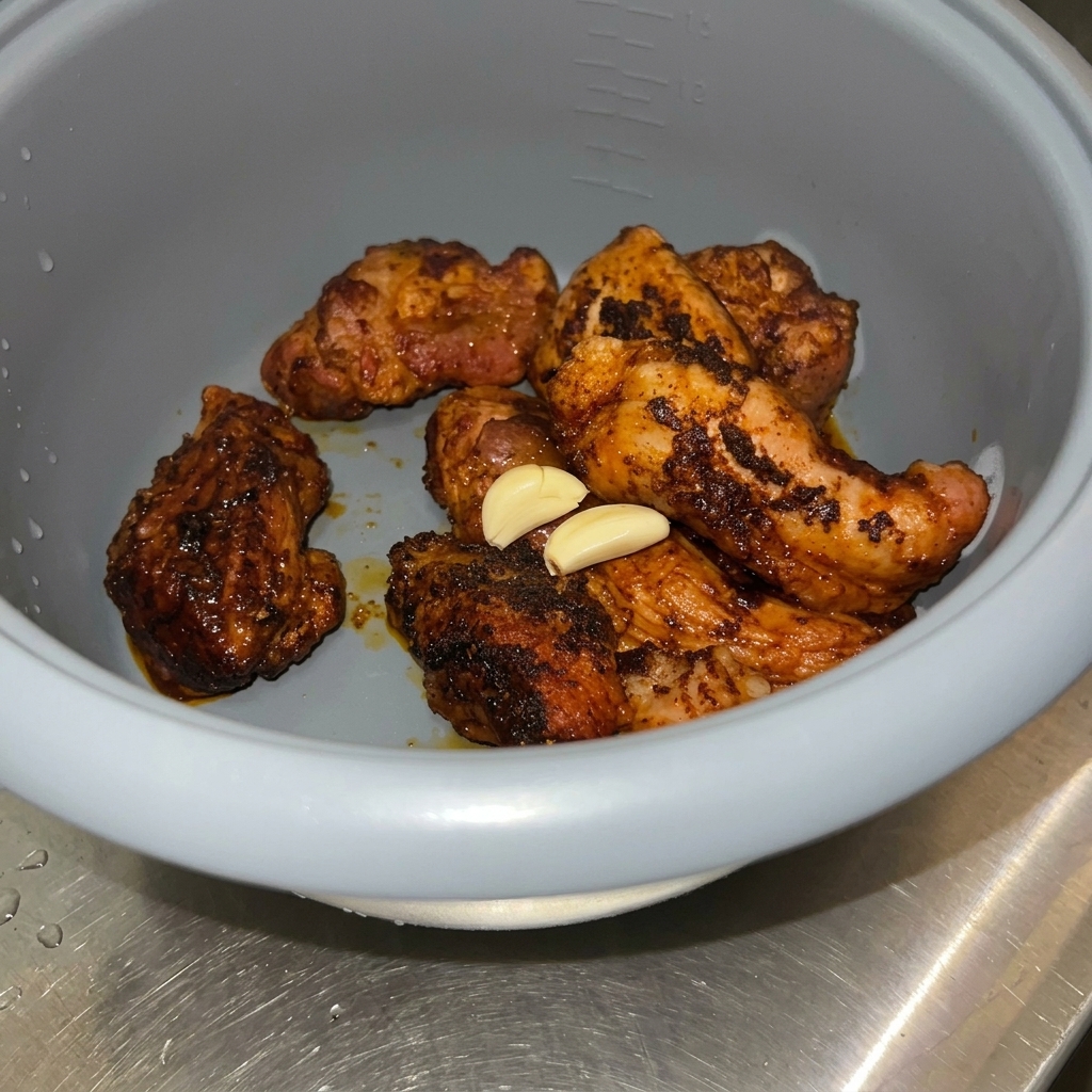 Peeled garlic cloves resting on top of browned pork chunks inside a grey rice cooker pot.
