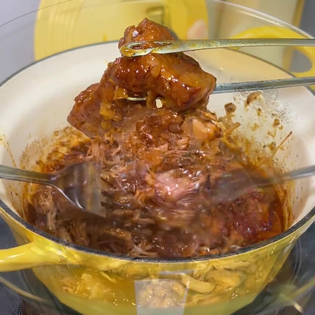 Tender, slow-cooked pork being shredded into fine strands with two metal forks inside a clear glass bowl.