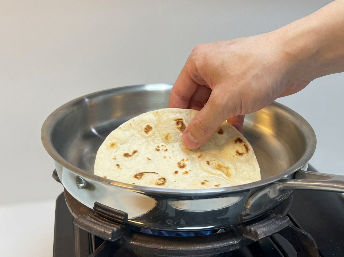 A hand placing a soft, thick flour tortilla onto a heated stainless steel pan.