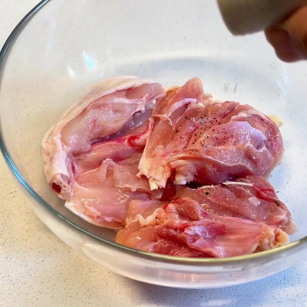 Raw chicken thighs in a clear glass bowl being seasoned with freshly ground black pepper from a grinder.
