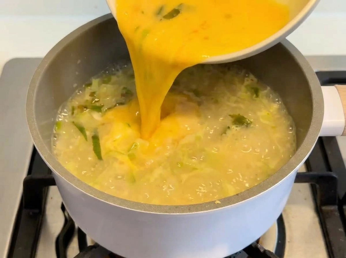 A vibrant yellow egg and lemon mixture being poured from a white bowl into a simmering pot of creamy soup.