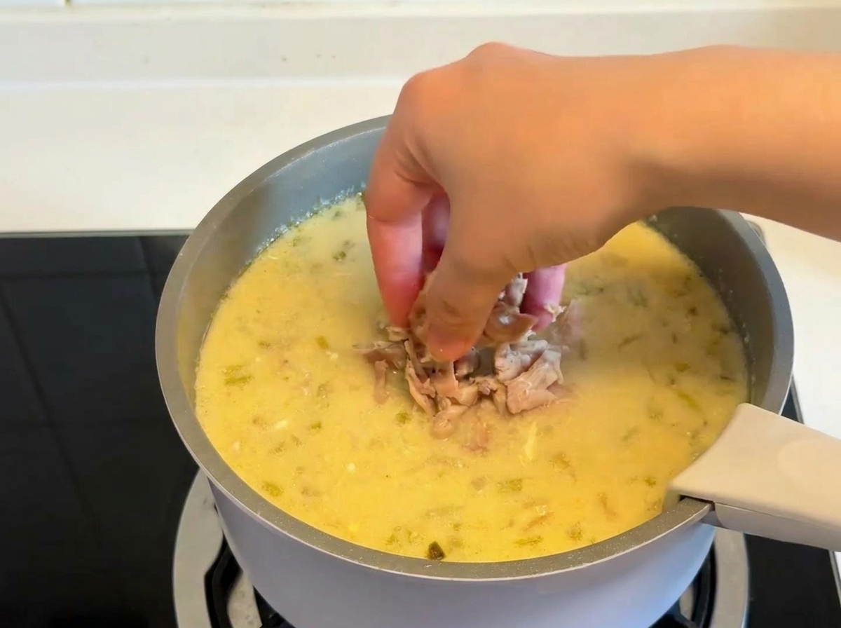 A hand dropping pieces of cooked, shredded chicken into a pot of thick, creamy lemon soup.
